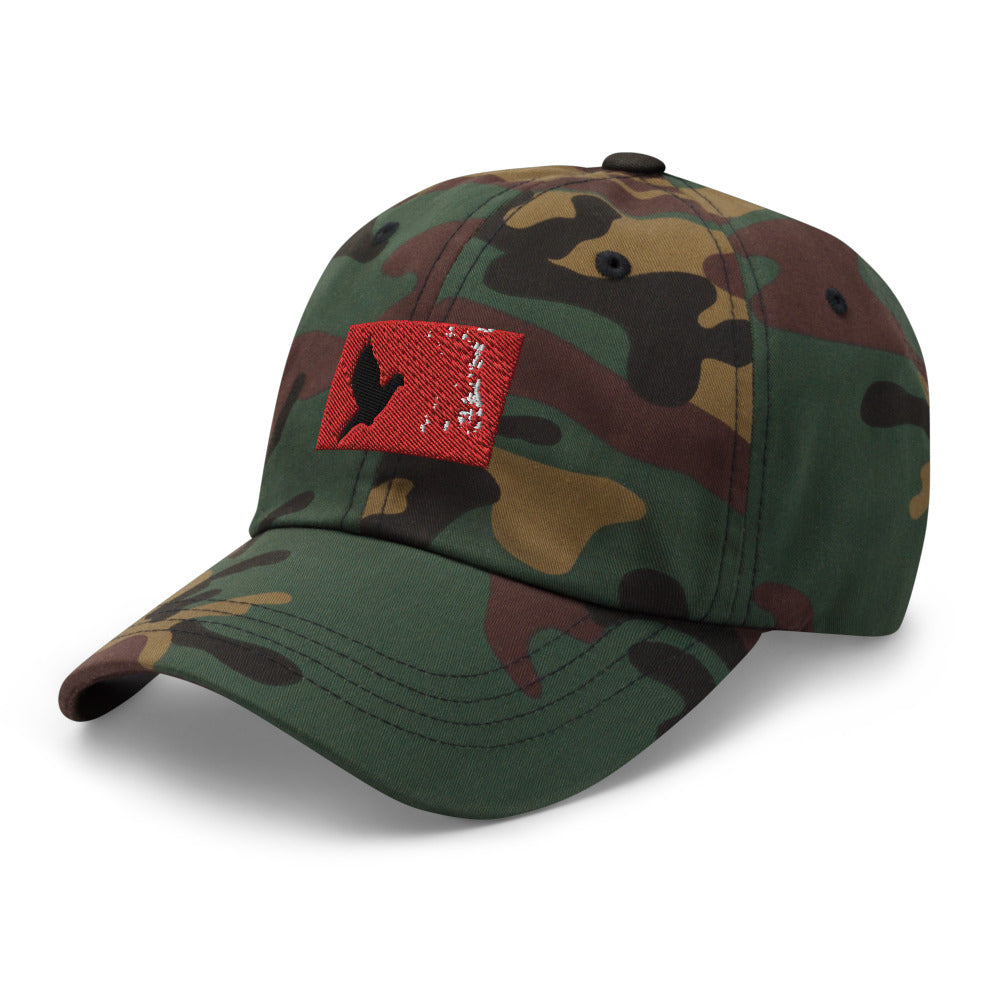 Camouflage dad hat with an embroidered red rectangle and a black pigeon flying on it. Hat is facing to it's right.