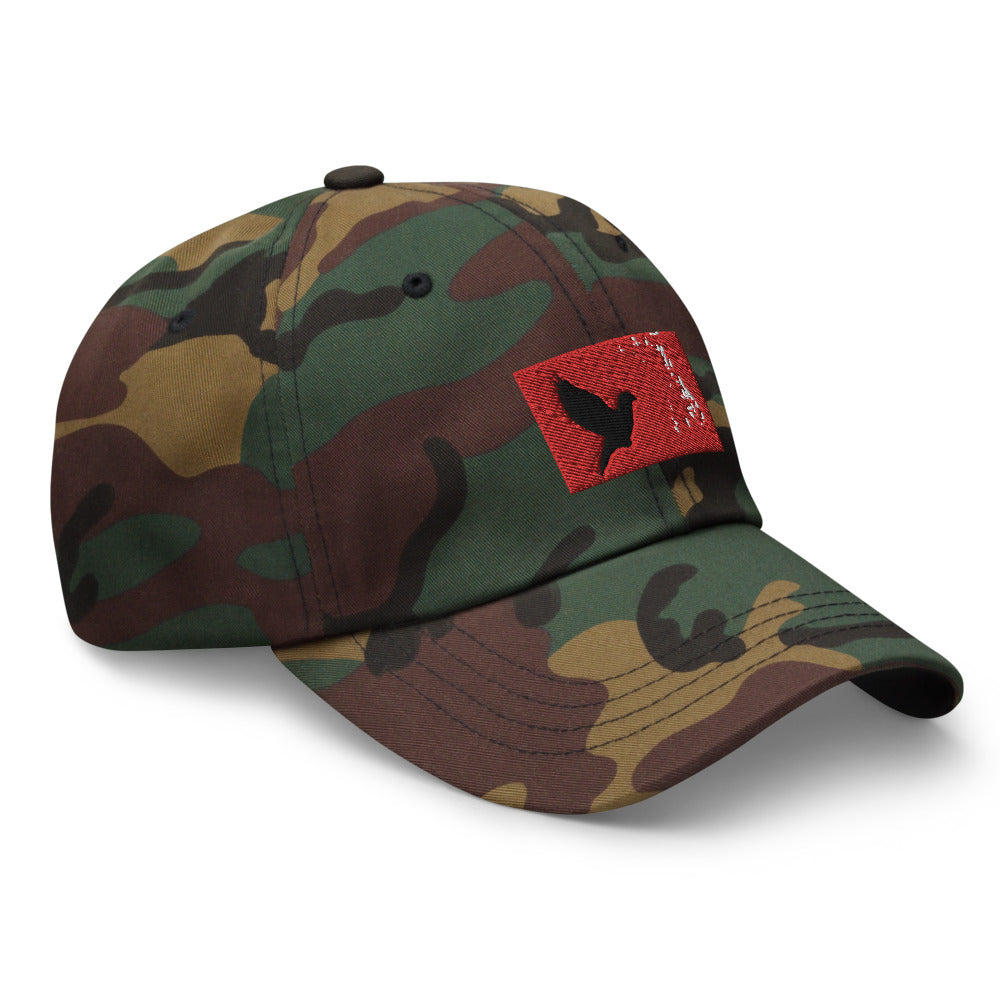 Camouflage dad hat with an embroidered red rectangle and a black pigeon flying on it. Hat is facing to it's left.