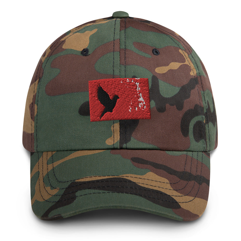 Camouflage dad hat with an embroidered red rectangle and a black pigeon flying on it. Close up pic.